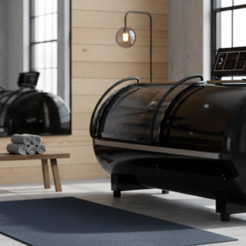 Black soft hyperbaric oxygen chamber in a minimalist biophilic wellness room with green living wall and sheer floor-to-ceiling curtains.
