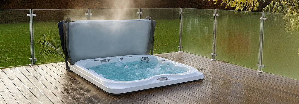 Steaming hot tub with raised cover on a wooden deck with glass railing overlooking a lush garden.