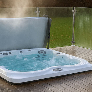 Steaming hot tub with raised cover on a wooden deck with glass railing overlooking a lush garden.