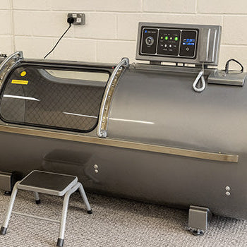 Hyperbaric oxygen chamber with digital control panel and step stool in a carpeted clinical room with white brick wall