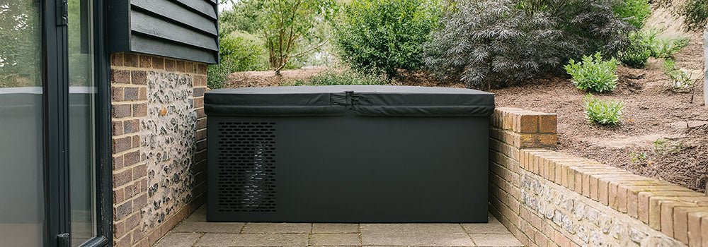 Black ice bath placed against a rustic house exterior, surrounded by stone paving and lush garden shrubs.