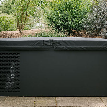 Black ice bath placed against a rustic house exterior, surrounded by stone paving and lush garden shrubs.
