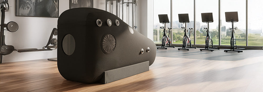 Inflatable hyperbaric oxygen chamber in a modern gym with hardwood floors, city views, and cardio equipment.