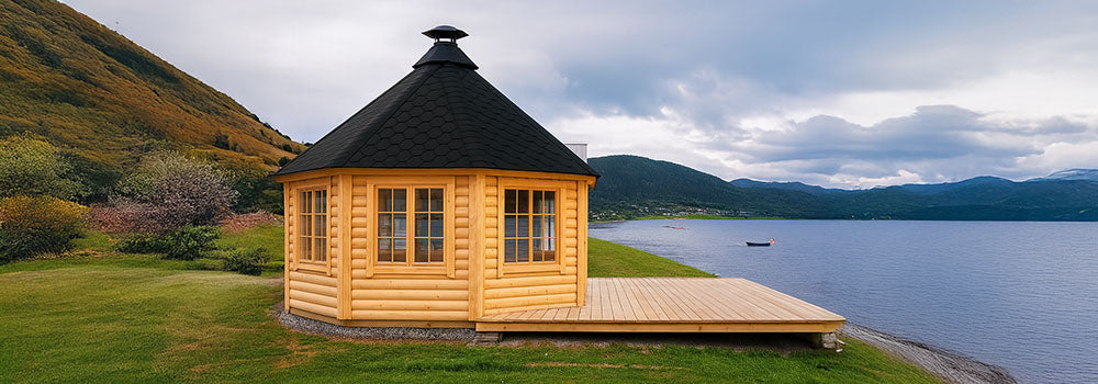 Octagonal wooden cabin with black roof on lakeside deck overlooking water and forested hills.