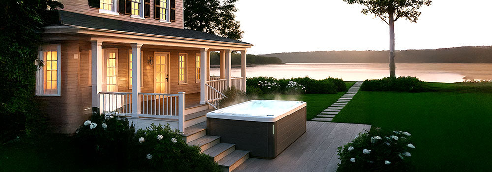 Steaming hot tub on lakeside deck at sunset beside a classic home with glowing windows.