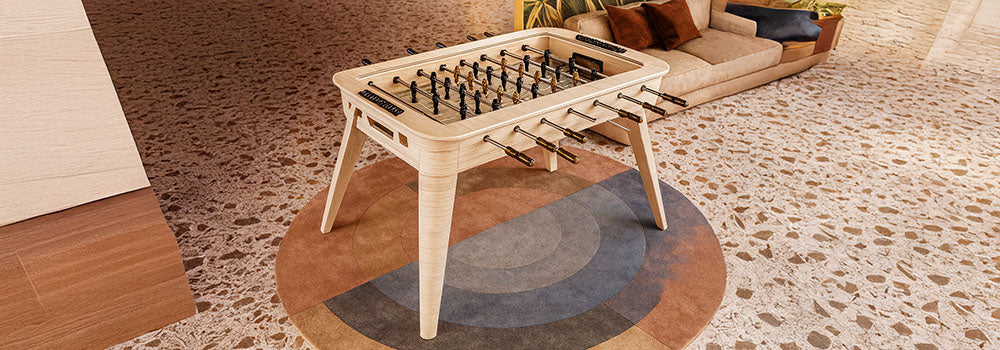 Sleek light wood foosball table in a mid-century modern living room with terrazzo flooring, tropical mural, and soft pendant lighting.