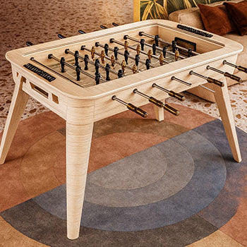 Sleek light wood foosball table in a mid-century modern living room with terrazzo flooring, tropical mural, and soft pendant lighting.