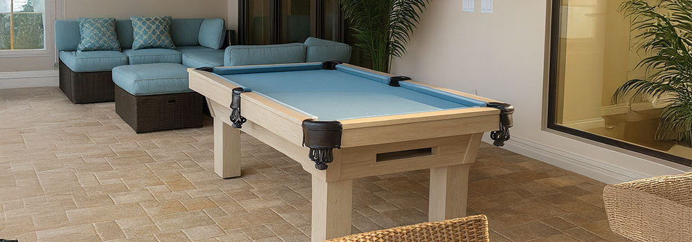 light wood pool table with sky-blue felt in a cozy indoor patio with wicker chairs, teal sectional, and potted plant.