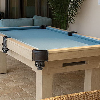 light wood pool table with sky-blue felt in a cozy indoor patio with wicker chairs, teal sectional, and potted plant.