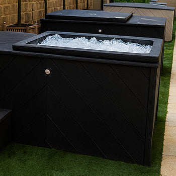 Row of black rectangular ice baths filled with ice in elegant outdoor courtyard with stone columns and greenery.