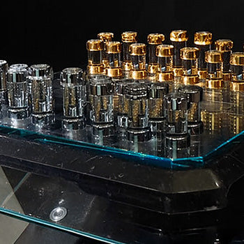 Luxury chess set with gold and silver pieces on a beveled glass board, displayed on a glossy black pedestal with glass shelving.