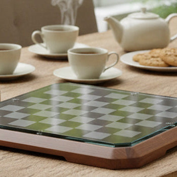 Modern glass chess set with gold and silver pieces placed beside a minimalist board on a wooden dining table, surrounded by tea set, books, and bright garden views.