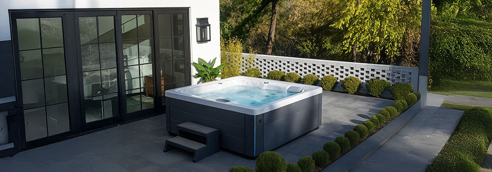 Square hot tub on a modern patio with white house exterior, black French doors, and landscaped greenery.