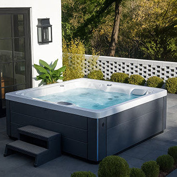 Square hot tub on a modern patio with white house exterior, black French doors, and landscaped greenery.