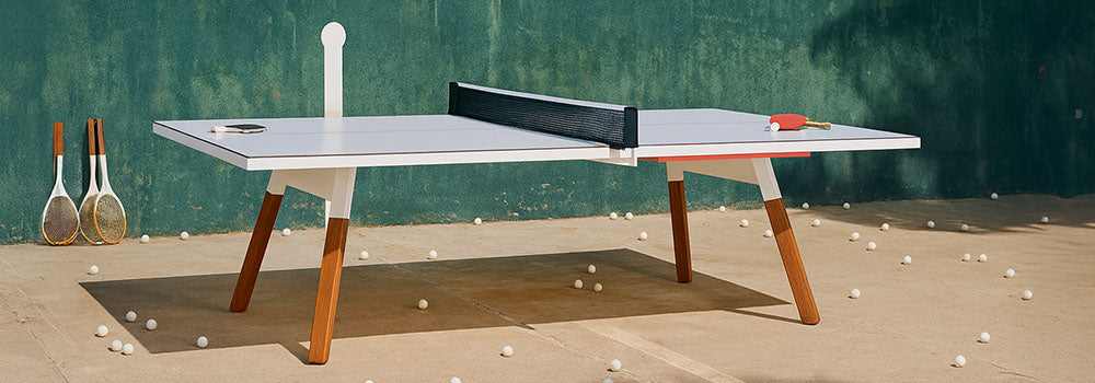 Modern outdoor table tennis table on a sunlit concrete patio with scattered ping pong balls and a green wall backdrop.