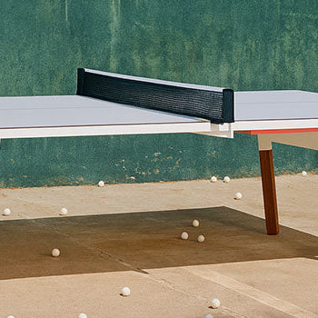 Modern outdoor table tennis table on a sunlit concrete patio with scattered ping pong balls and a green wall backdrop.
