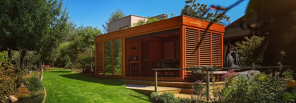 Sleek wooden garden room with slatted panels and glass walls, set on a manicured lawn surrounded by trees and flowering garden beds on a sunny day.