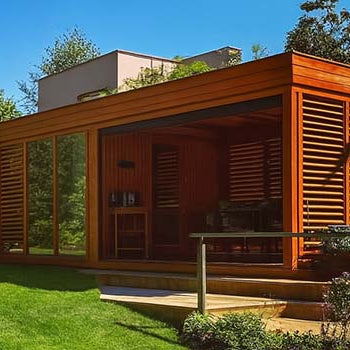 Sleek wooden garden room with slatted panels and glass walls, set on a manicured lawn surrounded by trees and flowering garden beds on a sunny day.