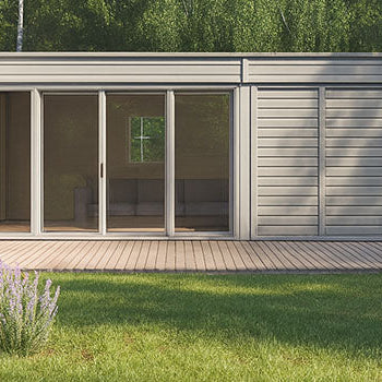 Modern L-shaped garden room with large glass doors, set in a lush green landscape with flowering plants and forest backdrop.