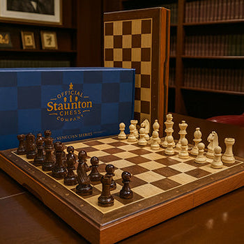 Official Staunton wooden chess set displayed in a refined library office, with rich wood tones, leather chair, book-lined shelves, and branded box in the background.
