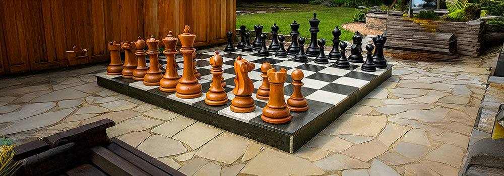 Oversized outdoor luxury garden chess set featuring large wooden and black pieces on a raised patio chessboard, surrounded by natural stone paving and landscaped greenery.