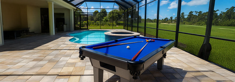 outdoor pool table with blue felt on a stone patio beside a screened-in swimming pool and hot tub, under a clear sunny sky.