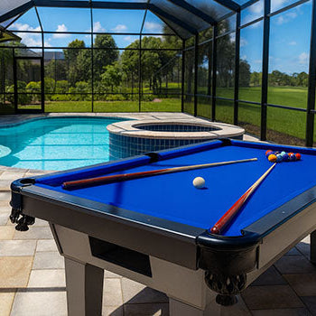 outdoor pool table with blue felt on a stone patio beside a screened-in swimming pool and hot tub, under a clear sunny sky.