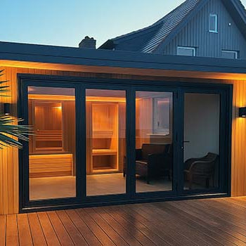 Modern outdoor sauna room with glass doors and wood cladding on a deck at twilight, attached to a traditional house.