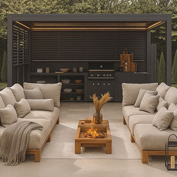Modern pergola with built-in outdoor kitchen, lounge seating, and fire pit surrounded by trees.