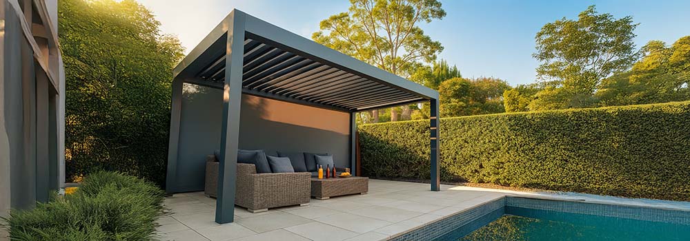 Modern dark grey pergola with wicker seating beside a pool, surrounded by greenery under clear skies.