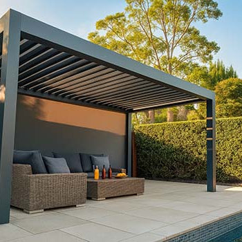 Modern dark grey pergola with wicker seating beside a pool, surrounded by greenery under clear skies.