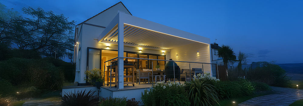 Illuminated white pergola with dining area and ceramic grill on an elevated patio outside a modern home at dusk.
