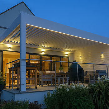 Illuminated white pergola with dining area and ceramic grill on an elevated patio outside a modern home at dusk.