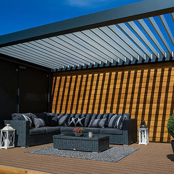 Stylish pergola with adjustable louvres over a dark wicker sofa set and rug, framed by lanterns and potted trees.