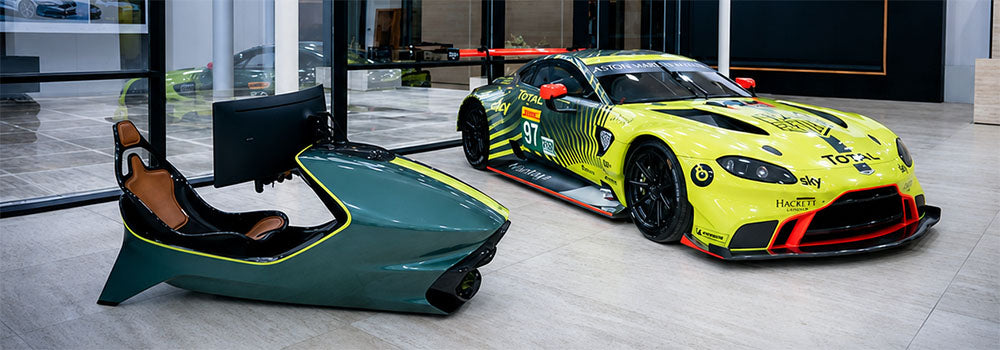 Premium racing simulator with sculpted cockpit and monitor displayed beside an Aston Martin race car in a luxury automotive showroom.
