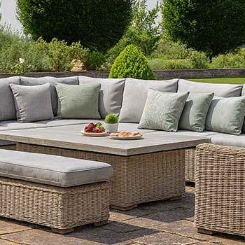 large rattan corner sofa set with grey cushions and bench on a paved garden patio surrounded by manicured lawns and tall trees.