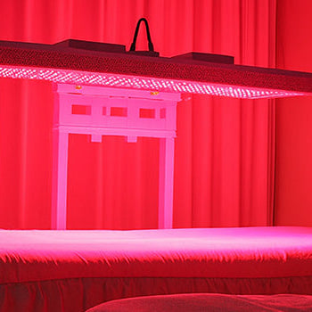 red light therapy panel suspended above a skirted treatment bed in a softly lit spa room with red curtains.