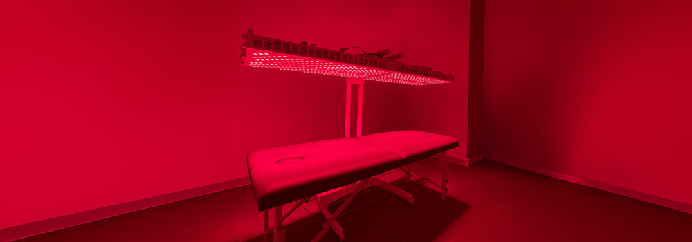 Large red light therapy panel glowing above cushioned treatment bed in minimalist red-lit room.