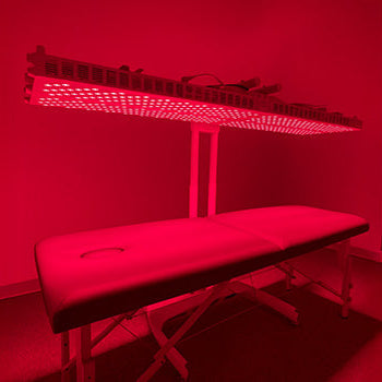 Large red light therapy panel glowing above cushioned treatment bed in minimalist red-lit room.