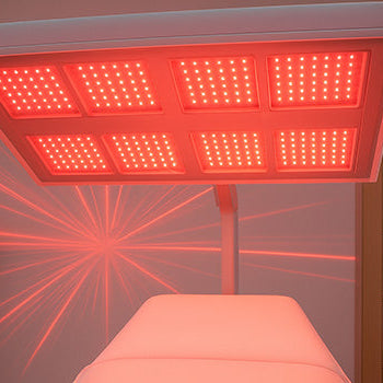 Overhead red light therapy panel illuminating a treatment bed in a modern spa room with skincare shelf and plant.