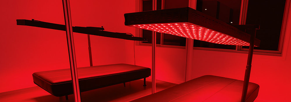red light therapy panel suspended above a black treatment bed in a mirrored room with glowing red ambiance.