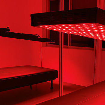 red light therapy panel suspended above a black treatment bed in a mirrored room with glowing red ambiance.