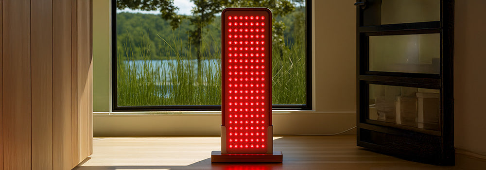 Red light therapy panel glowing beside a large window with lakeside and forest view.