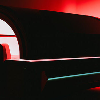 Futuristic red light therapy pod glowing in minimalist dark room with red wall reflections.