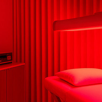 Minimalist red light therapy room with glowing treatment bed, curtain wall, and sideboard under ambient lighting.
