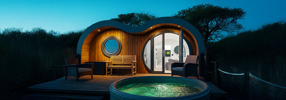 Glowing rotomoulded spa hot tub on a wooden deck outside a designer glamping pod at twilight.