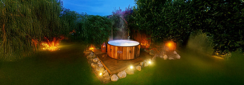 Steaming round wooden hot tub on deck with garden lighting at dusk.