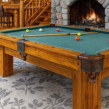rustic log cabin interior with a wooden pool table and stone fireplace under a wagon wheel chandelier.