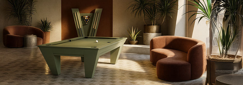 sage green pool table in a sunlit game room with curved velvet seating, tropical plants, and a V-shaped cue rack.