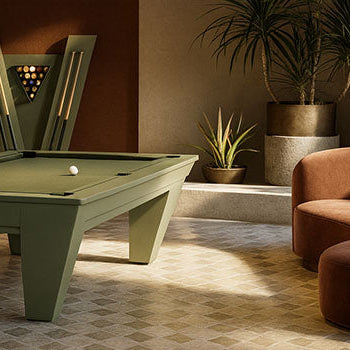sage green pool table in a sunlit game room with curved velvet seating, tropical plants, and a V-shaped cue rack.